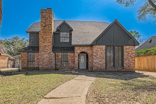 english style home with a front yard, fence, roof with shingles, a chimney, and brick siding