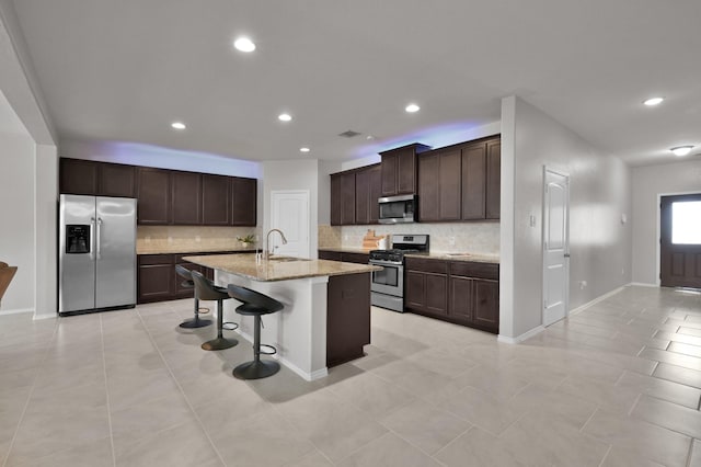 kitchen featuring light stone counters, a sink, dark brown cabinetry, appliances with stainless steel finishes, and a kitchen bar