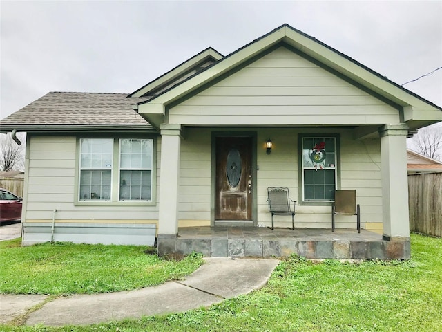 bungalow-style home with covered porch, a shingled roof, fence, and a front lawn