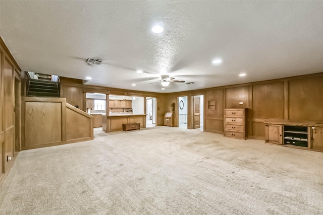 unfurnished living room featuring ceiling fan, a textured ceiling, a decorative wall, light carpet, and stairway