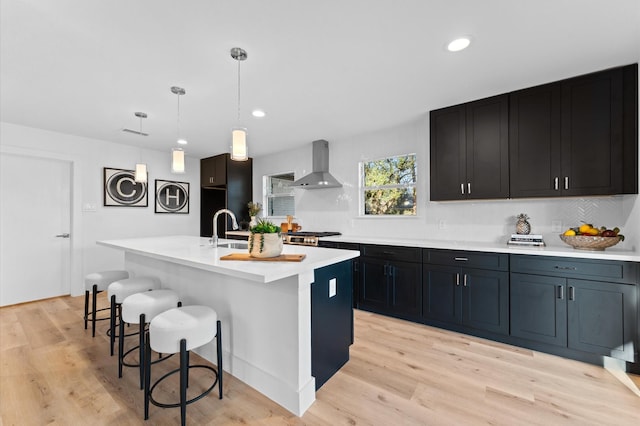 kitchen featuring wall chimney exhaust hood, light wood-style flooring, light countertops, a kitchen bar, and a sink