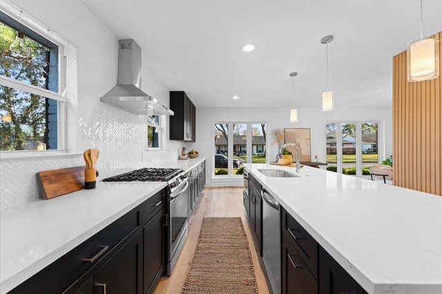 kitchen featuring appliances with stainless steel finishes, a sink, dark cabinetry, wall chimney range hood, and backsplash