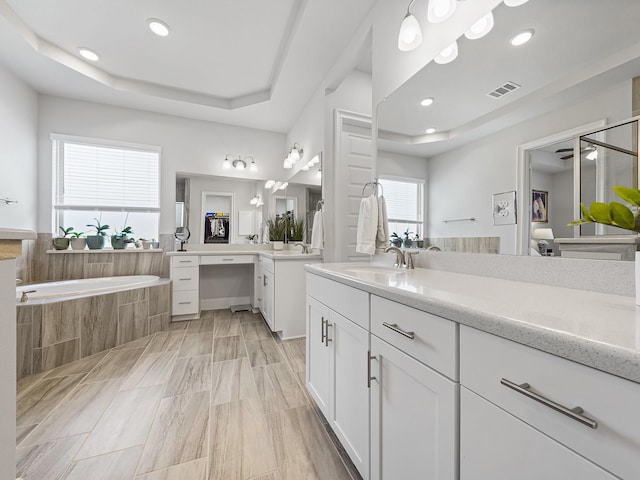 bathroom featuring visible vents, a tray ceiling, two vanities, a sink, and a bath