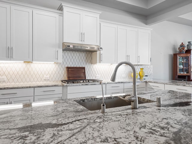 kitchen featuring light stone counters, stainless steel gas cooktop, white cabinets, under cabinet range hood, and tasteful backsplash