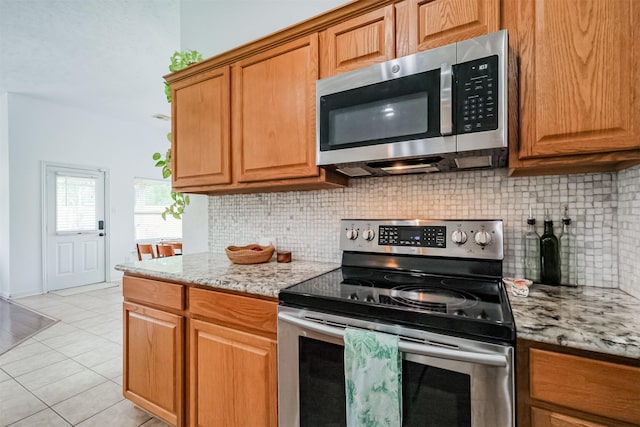 kitchen with light stone counters, brown cabinets, light tile patterned floors, backsplash, and appliances with stainless steel finishes