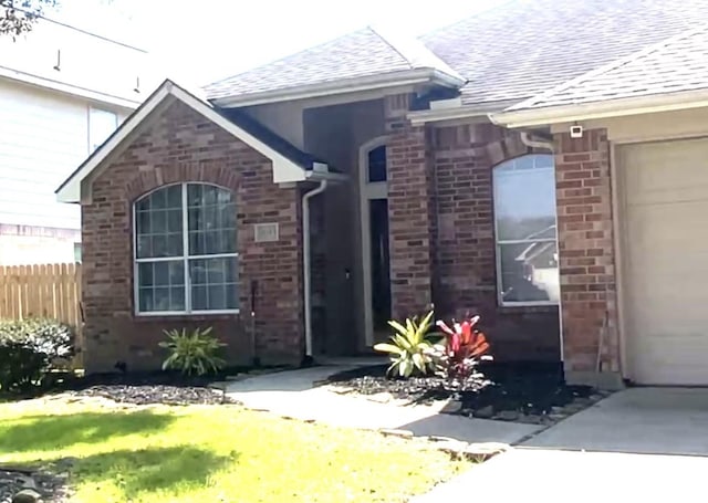 view of exterior entry featuring a garage, brick siding, roof with shingles, and fence