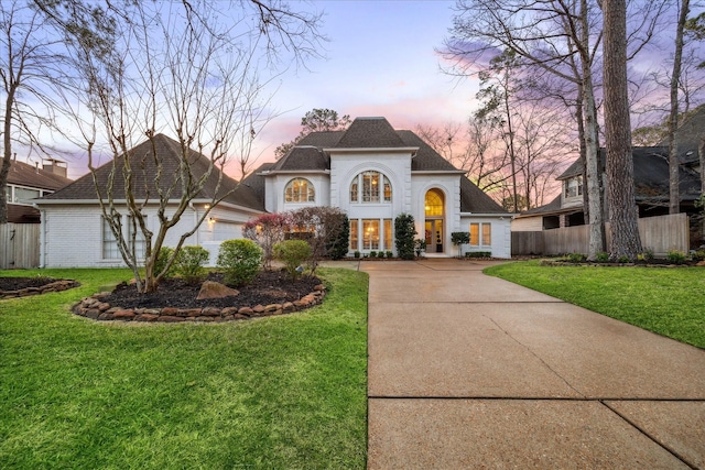 french country inspired facade featuring driveway, brick siding, fence, and a yard