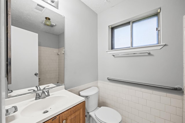 full bath featuring a textured ceiling, toilet, vanity, tile walls, and wainscoting