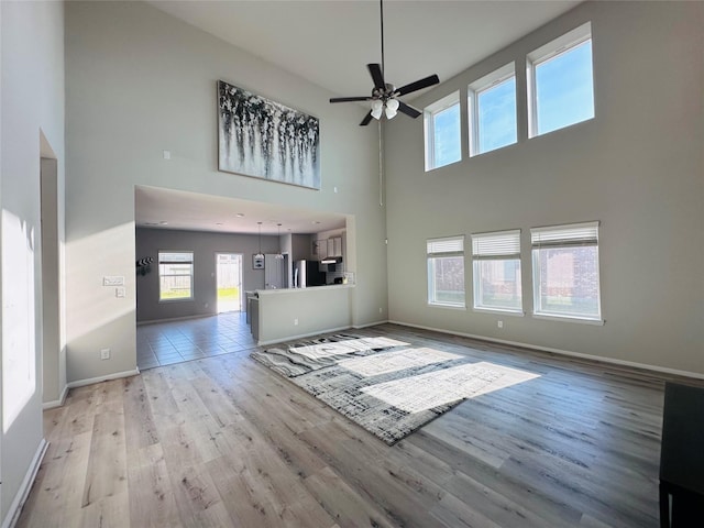 unfurnished living room featuring ceiling fan, light wood-type flooring, a towering ceiling, and baseboards