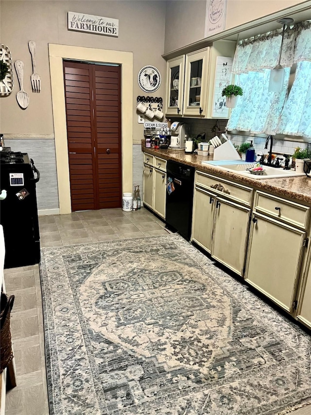 kitchen with light tile patterned floors, cream cabinets, a sink, black dishwasher, and glass insert cabinets