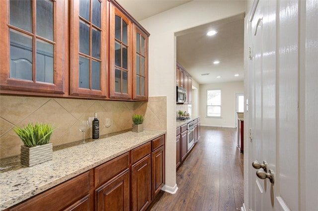 kitchen with dark wood-style flooring, tasteful backsplash, appliances with stainless steel finishes, glass insert cabinets, and light stone countertops