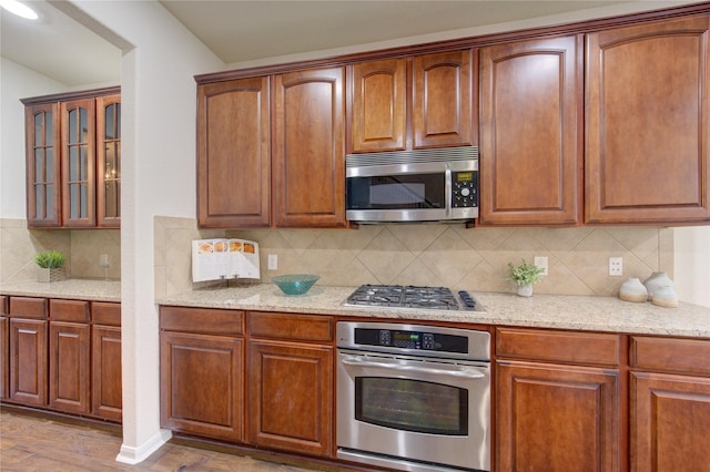 kitchen with tasteful backsplash, glass insert cabinets, wood finished floors, light stone countertops, and stainless steel appliances