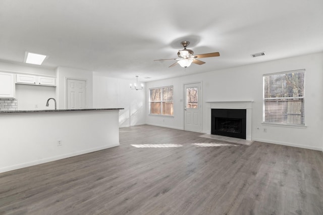 unfurnished living room with ceiling fan with notable chandelier, wood finished floors, a fireplace with flush hearth, visible vents, and baseboards