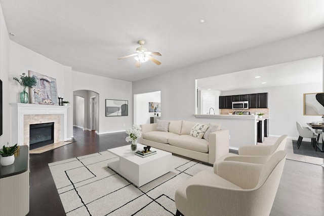 living room featuring light wood finished floors, arched walkways, baseboards, a fireplace with flush hearth, and ceiling fan