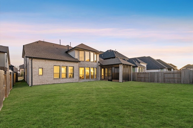 back of house at dusk featuring brick siding, roof with shingles, a lawn, a sunroom, and a fenced backyard