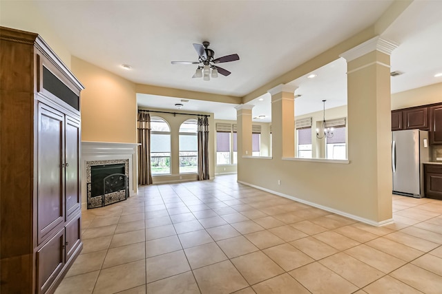 unfurnished living room with baseboards, ceiling fan with notable chandelier, ornate columns, a fireplace, and light tile patterned flooring