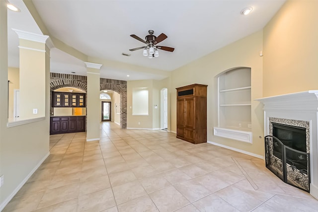 unfurnished living room with built in shelves, a ceiling fan, baseboards, a tiled fireplace, and ornate columns