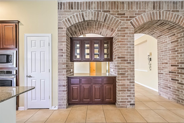 kitchen featuring arched walkways, light tile patterned floors, stainless steel appliances, glass insert cabinets, and dark stone counters