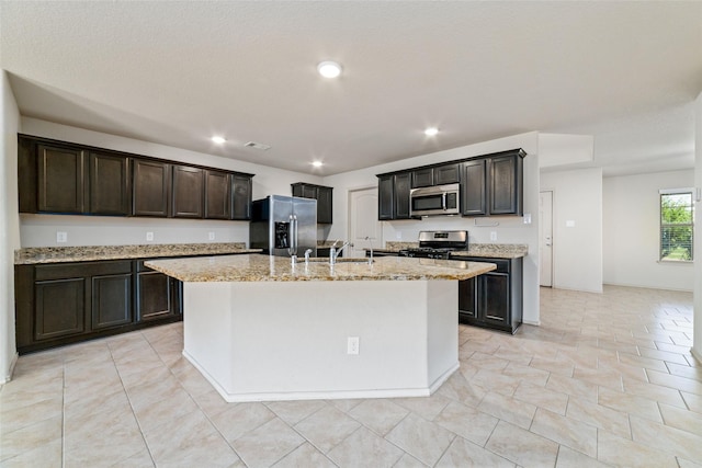 kitchen featuring a kitchen island with sink, recessed lighting, a sink, appliances with stainless steel finishes, and light stone countertops