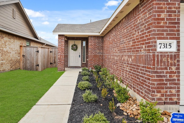 entrance to property with roof with shingles, brick siding, a lawn, and fence