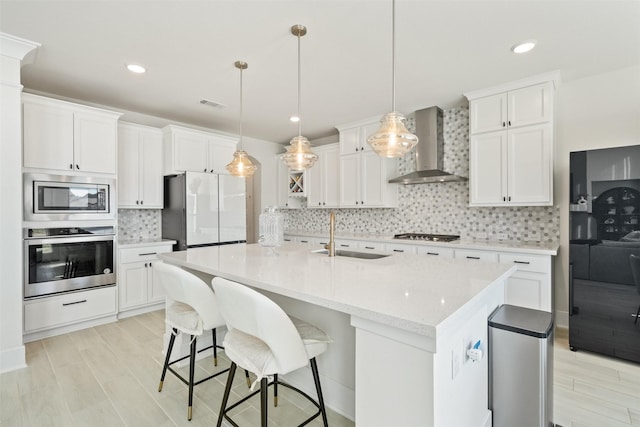 kitchen with stainless steel appliances, backsplash, white cabinetry, a sink, and wall chimney range hood