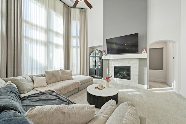 carpeted living area featuring baseboards, arched walkways, a ceiling fan, a high ceiling, and a fireplace