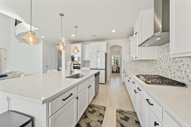 kitchen featuring arched walkways, stainless steel appliances, backsplash, a sink, and wall chimney exhaust hood