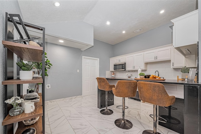 kitchen with dark countertops, stainless steel microwave, marble finish floor, vaulted ceiling, and a kitchen bar