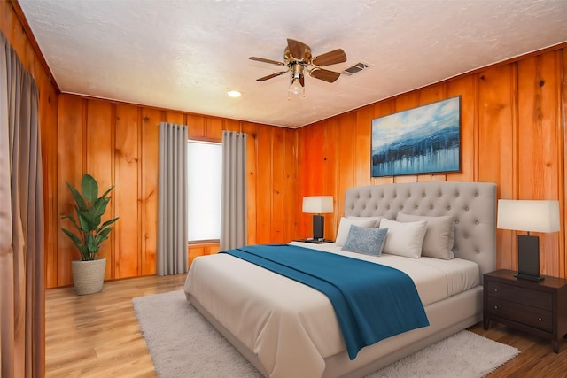 bedroom featuring visible vents, light wood-style floors, ceiling fan, a textured ceiling, and wooden walls