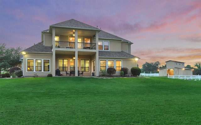 rear view of property featuring a shingled roof, a lawn, a balcony, ceiling fan, and fence