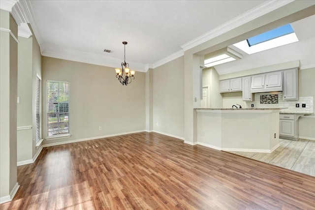 unfurnished living room with a skylight, a notable chandelier, crown molding, light wood-type flooring, and baseboards