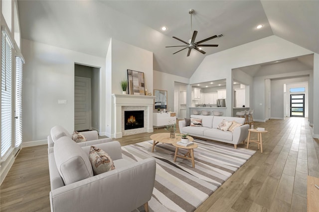 living room with high vaulted ceiling, light wood-style flooring, a warm lit fireplace, baseboards, and ceiling fan