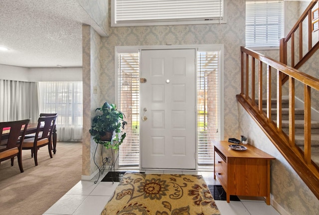 foyer entrance with a textured ceiling, light tile patterned floors, wallpapered walls, stairs, and light carpet