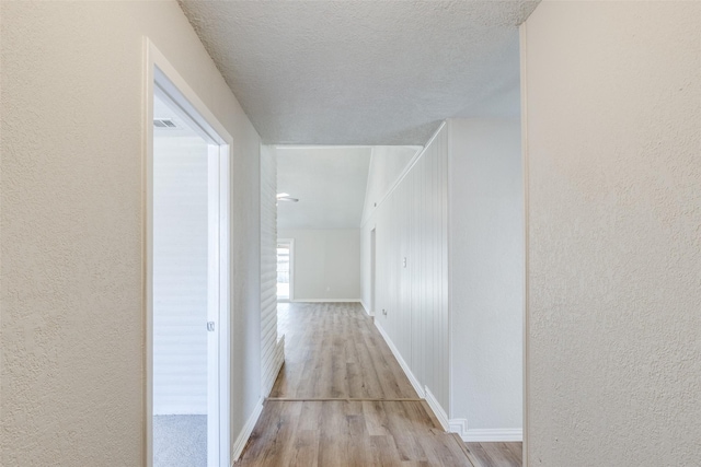 hall with a textured ceiling, a textured wall, wood finished floors, and baseboards