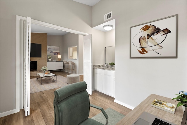 home office featuring light wood finished floors, a brick fireplace, visible vents, and baseboards