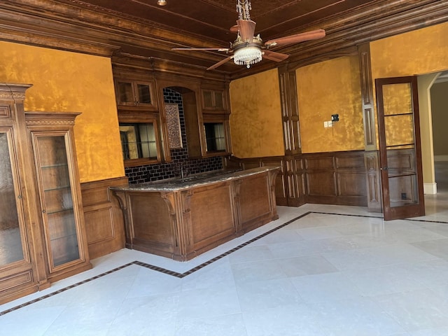 11117 Beinhorn Rd, Houston, TX 77024 - kitchen featuring a ceiling fan, glass insert cabinets, wainscoting, brown cabinets, and dark stone counters