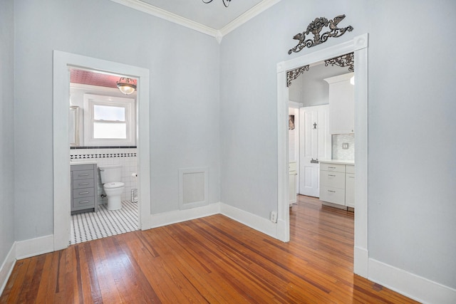 unfurnished bedroom with tile walls, ornamental molding, ensuite bath, baseboards, and hardwood / wood-style flooring