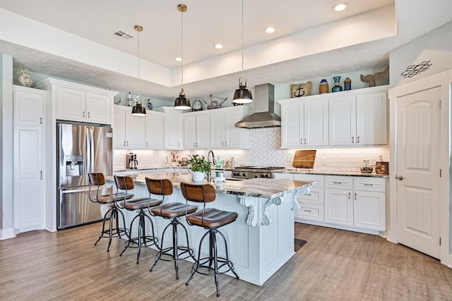 kitchen with visible vents, a raised ceiling, stove, wall chimney range hood, and stainless steel refrigerator with ice dispenser