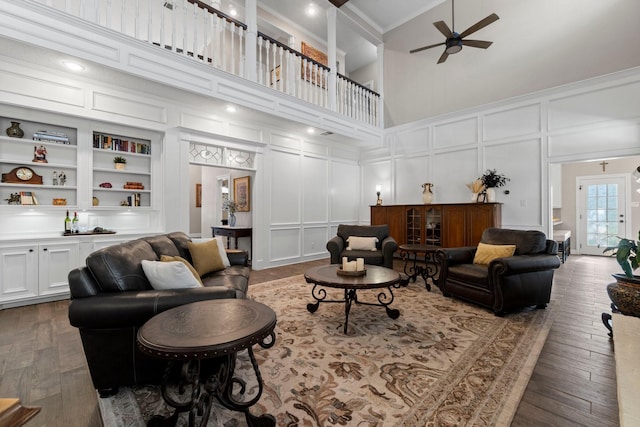 living room with ceiling fan, wood-type flooring, a high ceiling, built in shelves, and a decorative wall