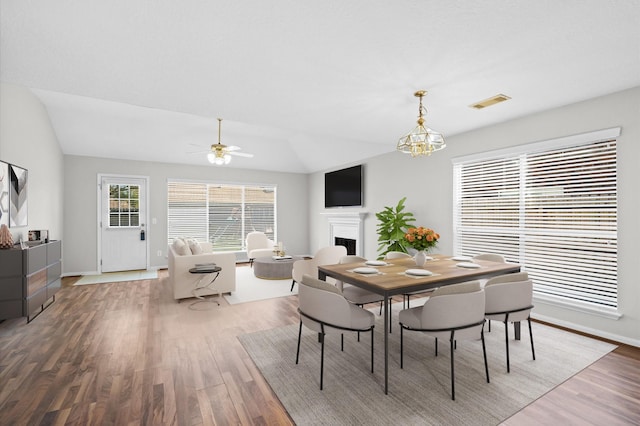 dining area with a fireplace, vaulted ceiling, wood finished floors, and ceiling fan with notable chandelier