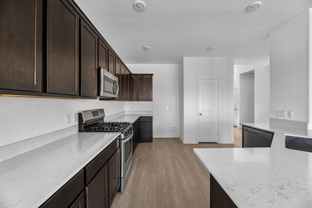 kitchen featuring a textured ceiling, light wood-style floors, dark brown cabinets, appliances with stainless steel finishes, and light stone countertops