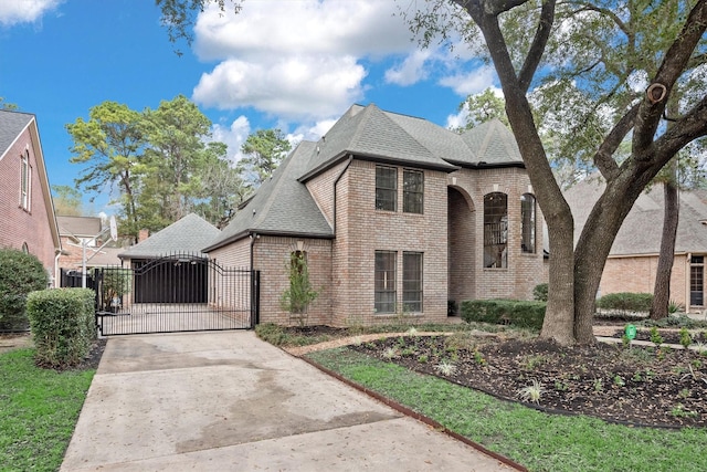 french country home featuring a shingled roof, a gate, concrete driveway, and brick siding