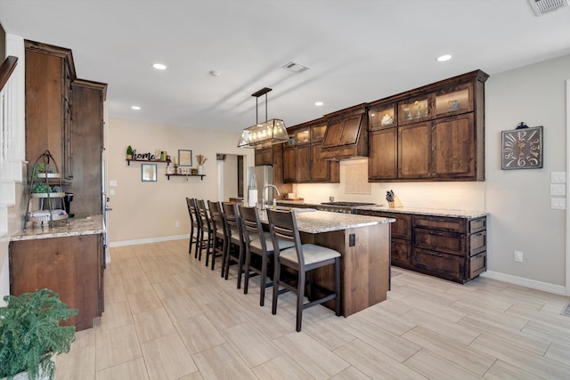 kitchen with visible vents, custom range hood, a kitchen island with sink, stainless steel appliances, and dark brown cabinets