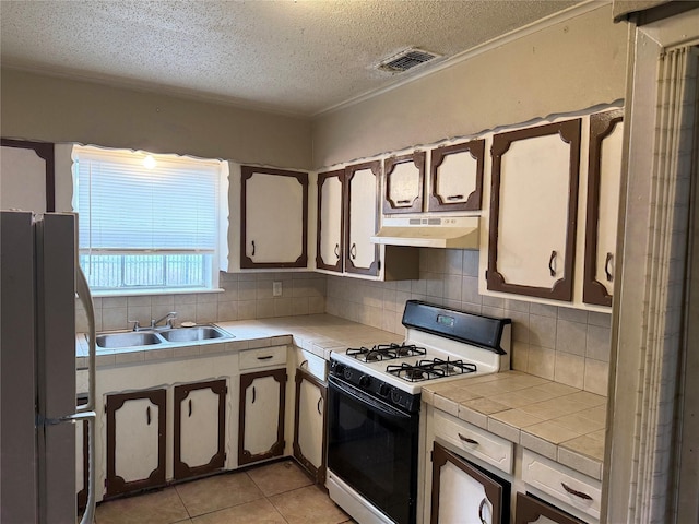 kitchen with tile counters, freestanding refrigerator, gas range oven, under cabinet range hood, and a sink