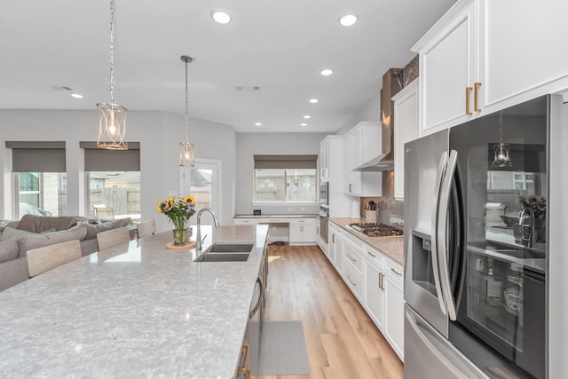 kitchen featuring white cabinets, a sink, light wood-style floors, wall chimney exhaust hood, and appliances with stainless steel finishes