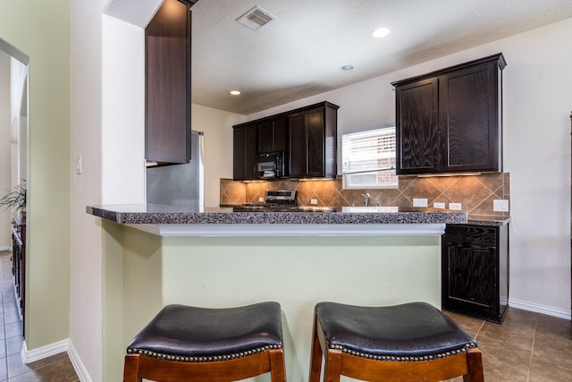 kitchen with stainless steel appliances, tile patterned flooring, dark brown cabinets, a kitchen breakfast bar, and backsplash