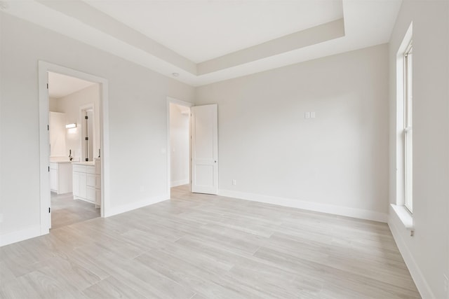 unfurnished bedroom featuring a tray ceiling, light wood-type flooring, ensuite bath, and baseboards