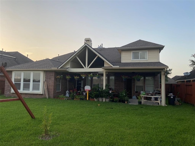 back of house at dusk with a yard, brick siding, a shingled roof, and fence