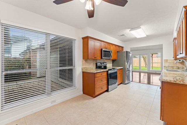 kitchen with brown cabinetry, decorative backsplash, light stone counters, stainless steel appliances, and a sink