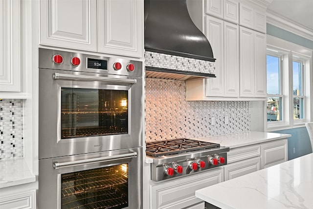 kitchen featuring decorative backsplash, appliances with stainless steel finishes, light stone countertops, premium range hood, and white cabinetry